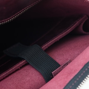 Puede incluir: Primer plano de un estuche de cuero burdeos con cremallera negra y banda el&aacute;stica. El interior revela un bolsillo y un forro oscuro. El estuche est&aacute; dise&ntilde;ado para guardar objetos de forma segura.
