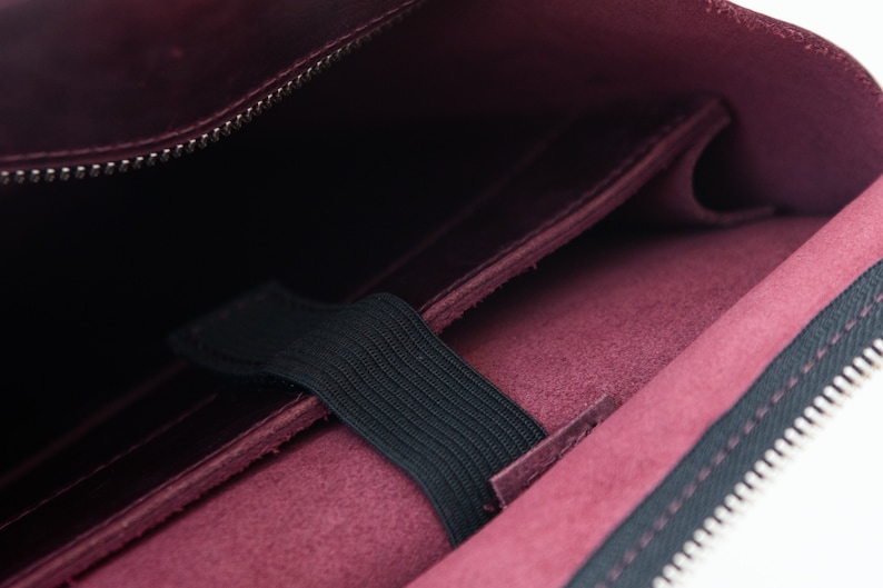 Puede incluir: Primer plano de un estuche de cuero burdeos con cremallera negra y banda el&aacute;stica. El interior revela un bolsillo y un forro oscuro. El estuche est&aacute; dise&ntilde;ado para guardar objetos de forma segura.