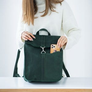 May include: A dark green leather backpack with a top handle and a front pocket. The backpack has a flap closure with a silver-coloured clasp. A smartphone is visible in the front pocket. The bag is held by a person wearing a white jumper.