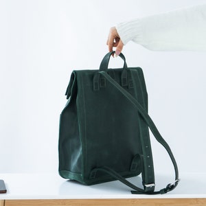 May include: A dark green backpack with a rectangular shape and a flap closure. The backpack has adjustable straps and a top handle. The bag is made of a textured material and is on a white surface.