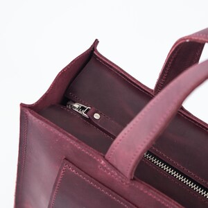 Puede incluir: Primer plano de un bolso tote de cuero burdeos con cierre de cremallera plateada. El bolso tiene dos asas y forma rectangular. El cuero parece texturizado y el bolso est&aacute; sobre un fondo blanco.