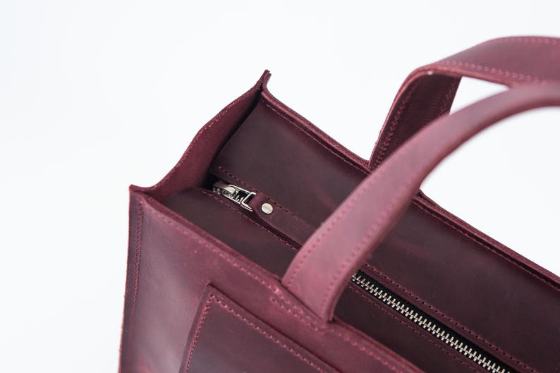 Puede incluir: Primer plano de un bolso tote de cuero burdeos con cierre de cremallera plateada. El bolso tiene dos asas y forma rectangular. El cuero parece texturizado y el bolso est&aacute; sobre un fondo blanco.