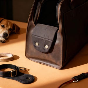 May include: A brown leather pet carrier with a dark brown fabric pocket secured by two gold-colored buttons. A black and gold dog collar and a beige and brown collar are on the table. A small dog is in the background.