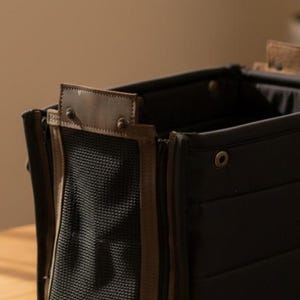 May include: Close-up of a black pet carrier with brown leather accents. The carrier features a mesh panel and a zipper closure. The top has a brown leather handle and is secured with metal rivets. The carrier is designed for pet travel.