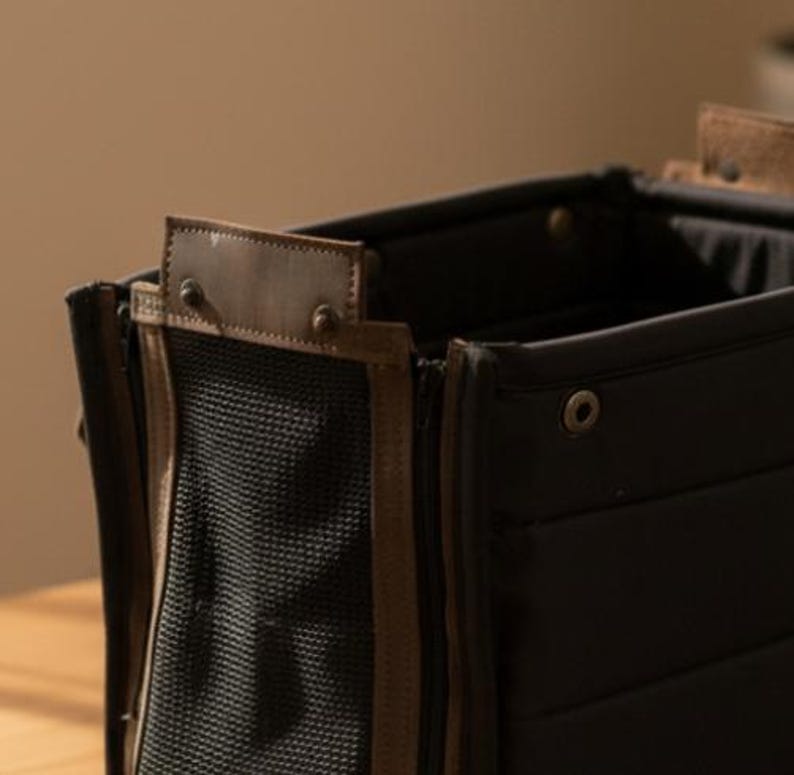 May include: Close-up of a black pet carrier with brown leather accents. The carrier features a mesh panel and a zipper closure. The top has a brown leather handle and is secured with metal rivets. The carrier is designed for pet travel.