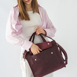 Puede incluir: Bolso bandolera de cuero burdeos con bolsillo con solapa y correa para el hombro. El bolso tiene herrajes plateados y un dise&ntilde;o estructurado. El bolso lo sostiene una persona que lleva una camisa rosa claro y pantalones blancos.