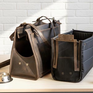 May include: Two pet carriers, one brown leather and one black fabric, sit on a light wood surface. A stainless steel pet bowl with paw prints and a colorful rope toy are also present. The leather carrier has a handle and shoulder strap.