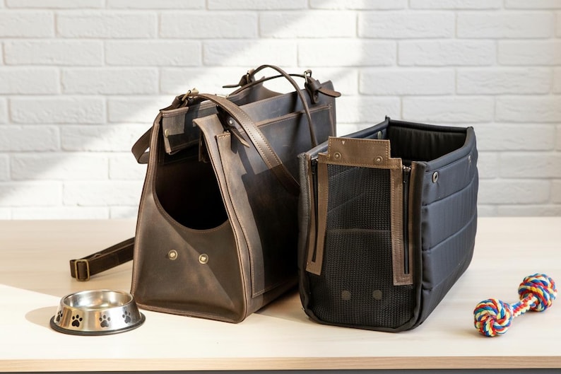 May include: Two pet carriers, one brown leather and one black fabric, sit on a light wood surface. A stainless steel pet bowl with paw prints and a colorful rope toy are also present. The leather carrier has a handle and shoulder strap.