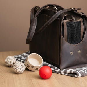 May include: A brown leather pet carrier with a mesh window, handles, and a shoulder strap. Beside it are two rope toys, a ceramic bowl with paw prints, and a red ball, all on a striped blanket.