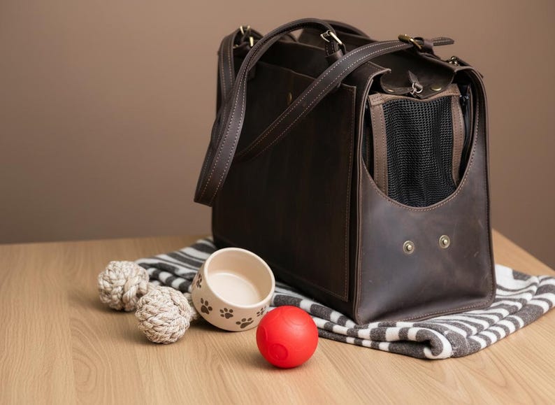 May include: A brown leather pet carrier with a mesh window, handles, and a shoulder strap. Beside it are two rope toys, a ceramic bowl with paw prints, and a red ball, all on a striped blanket.