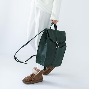 Puede incluir: Una mochila de cuero verde oscuro con cierre de solapa y un bolsillo delantero. La mochila tiene correas ajustables y un asa superior. La persona lleva pantalones blancos y un suéter blanco.