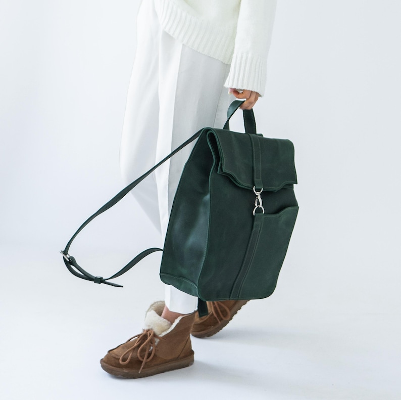 May include: A dark green leather backpack with a flap closure and a front pocket. The backpack has adjustable straps and a top handle. The bag is being carried by someone wearing white trousers and a white jumper.