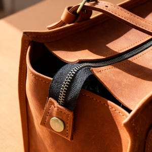 May include: Close-up of a brown leather tote bag with a black zipper. The bag has a handle and a brass-colored button. The leather has a natural, textured appearance. The bag is designed for carrying items.