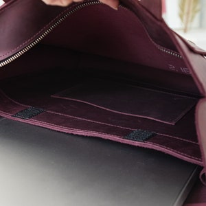 Puede incluir: Vista abierta de un bolso para port&aacute;til de cuero burdeos con cremallera y herrajes plateados. El interior revela un port&aacute;til, un peque&ntilde;o bolsillo y un sello de fecha. El bolso est&aacute; dise&ntilde;ado para llevar un port&aacute;til y otros elementos esenciales.