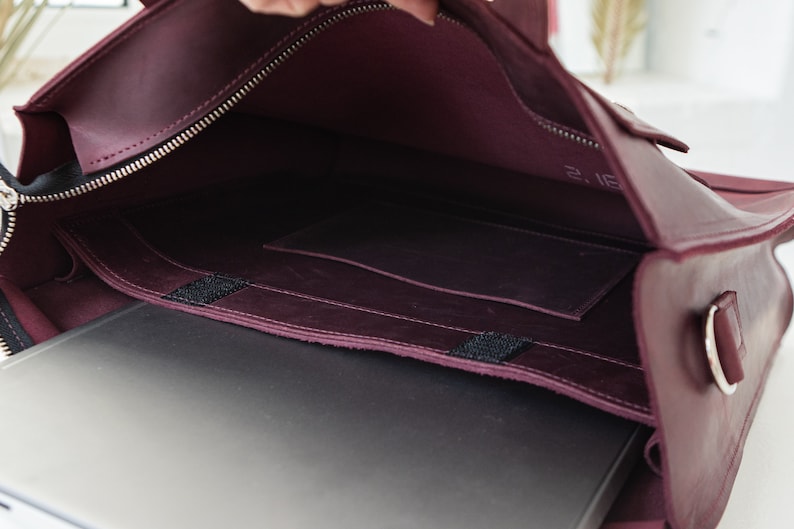 Puede incluir: Vista abierta de un bolso para port&aacute;til de cuero burdeos con cremallera y herrajes plateados. El interior revela un port&aacute;til, un peque&ntilde;o bolsillo y un sello de fecha. El bolso est&aacute; dise&ntilde;ado para llevar un port&aacute;til y otros elementos esenciales.