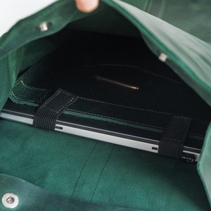 May include: A dark green leather laptop bag with a flap closure. The bag is open, revealing a silver laptop secured by a black elastic strap. The bag is made of a soft, textured leather.