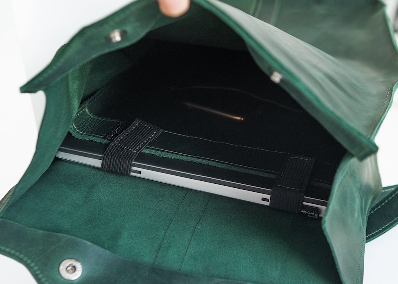 May include: A dark green leather laptop bag with a flap closure. The bag is open, revealing a silver laptop secured by a black elastic strap. The bag is made of a soft, textured leather.