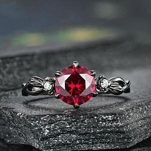 以下が含まれることがあります： 黒色のリングに、大きな赤い宝石と、両側に小さな白い宝石が2つ付いています。リングは暗い灰色の石の上に置かれています。