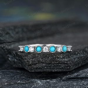 May include: A silver ring featuring a row of round, bright turquoise stones alternating with smaller, clear gemstones. The ring is set on a dark, textured surface, showcasing the jewelry's sparkle and design.