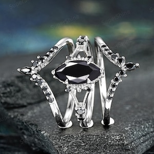 May include: A silver ring set with a large black marquise-shaped gemstone surrounded by smaller black gemstones. The ring is made up of three bands that are intertwined.
