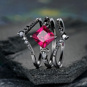 May include: A three-piece ring set featuring a central ring with a large, square, bright pink gemstone. The set includes two additional rings, one with a black stone accent and the other with a black stone accent. The rings are set on a dark gray surface.