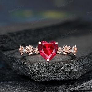 Peut inclure: Une bague en or rose ornée d'une pierre rouge vif en forme de cœur. La monture est ornée de motifs floraux et de petites pierres. La bague est posée sur un fond sombre et texturé.