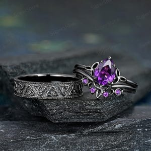 Könnte beinhalten: Zwei schwarze Ringe. Ein Ring hat ein Band mit keltischem Knotenmuster. Der andere Ring hat einen birnenförmigen lila Edelstein und kleinere lila Edelsteine. Die Ringe liegen auf einer dunklen, strukturierten Oberfläche.