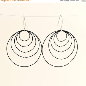 May include: A pair of black hoop earrings with a layered, circular design. Each earring features multiple concentric circles, creating a bold, geometric look. The earrings hang from silver-colored ear wires, offering a modern and minimalist aesthetic.