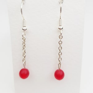 May include: A pair of drop earrings with a silver chain and a small, round, red bead. The earrings have a simple hook design for pierced ears. The background is white.