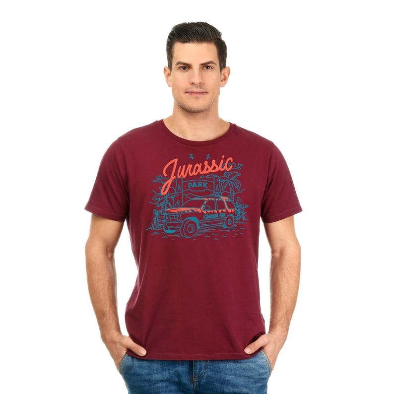 Pu&ograve; includere: Una t-shirt color granata con una stampa grafica di una jeep che guida attraverso una giungla con il testo "Jurassic Park" in un font blu e arancione.