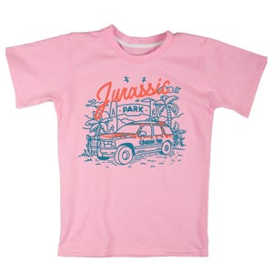 Pu&ograve; includere: Maglietta rosa con una grafica blu e arancione di una jeep che guida attraverso una giungla con il testo "Jurassic Park" sul lato della jeep.