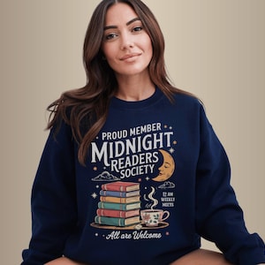 Könnte beinhalten: Marineblaues Sweatshirt mit dem Text "Proud Member Midnight Readers Society". Das Design umfasst einen Stapel Bücher, einen Mond und eine Tasse Tee. Der Satz "All are Welcome" ist ebenfalls auf dem Sweatshirt aufgedruckt.
