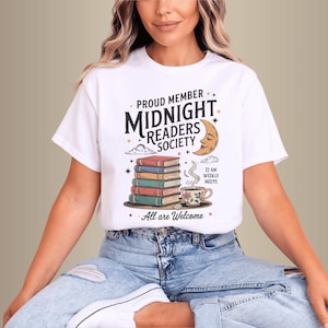 Pode incluir: T-shirt branca com a frase "Proud Member Midnight Readers Society", uma pilha de livros, uma lua crescente e uma chávena de chá. A t-shirt é de estilo casual e confortável, ideal para os amantes de livros e leitura.