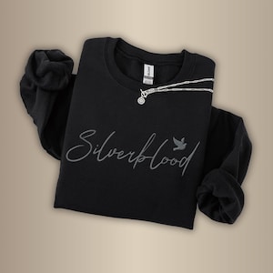 May include: Black crew neck sweatshirt with the text "Silverblood" in a script font and a small bird graphic. A silver necklace with a circular pendant is draped across the shirt. The word "Black" is in a black circle in the upper left corner.
