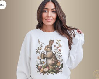 Può includere: Felpa bianca con un coniglio marrone che tiene un annaffiatoio circondato da fiori e foglie. Il coniglio è al centro del design.