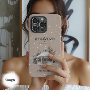 Peut inclure: Coque de téléphone beige avec une scène hivernale représentant un gazebo et de la neige qui tombe. La coque porte l'inscription "STARS HOLLOW FOUNDED 1779" et "i smell snow...". La coque est étiquetée "Tough".