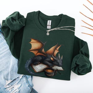 Cozy sweatshirt with cute black dragon sleeping on book on dragonkind with mug of coffee with moon phase pattern. Fourth wing Tairn Andarna dragons bookish booktok bookstagram  fantasy fan reader romantasy coffee lover gift idea soft warm sweater GOT