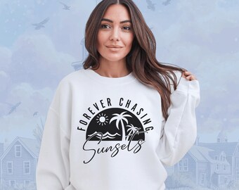 May include: White sweatshirt with the text "FOREVER CHASING SUNSETS" in a black, circular design featuring a sun, palm trees, and waves. The sweatshirt is a casual, long-sleeved garment.