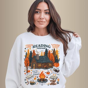 Pode incluir: Camisola branca com um gráfico colorido de uma cabana na floresta, uma fogueira e as palavras "Reading Retreat". O design inclui folhagem de outono, abóboras e livros, criando um tema outonal e acolhedor.