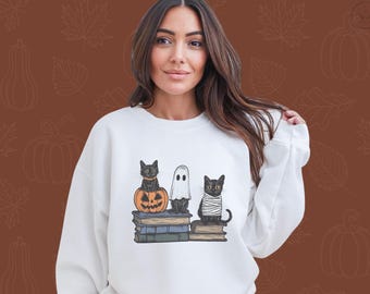以下が含まれることがあります： ハロウィーンをテーマにしたグラフィックが特徴の白いスウェットシャツ。デザインには、カボチャに乗った黒猫、幽霊、包帯を巻いた猫が描かれており、すべて本の上に座っています。背景は、葉とカボチャの模様が入った茶色です。