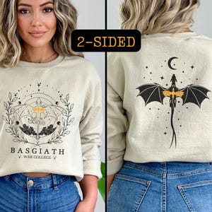 Könnte beinhalten: Ein doppelseitiges cremefarbenes Sweatshirt mit einem grafischen Design auf der Vorderseite und einem Drachen-Design auf der Rückseite. Das Design auf der Vorderseite zeigt eine Illustration zum Thema Himmel mit dem Text "Basgiath *War College*" darunter. Das Design auf der Rückseite zeigt einen schwarzen Drachen mit gelben Akzenten, der vor einem sternenklaren Nachthimmel fliegt.