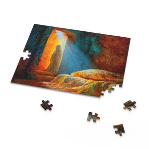 May include: A partially assembled jigsaw puzzle showing a religious scene. The artwork features a figure in a white robe standing in a bright doorway, with a tomb in the foreground. Several puzzle pieces are scattered around the main image.