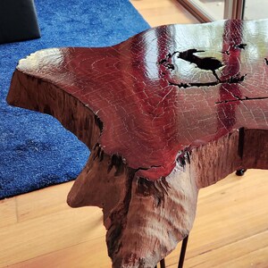 May include: A handcrafted wooden side table with a natural shape. The tabletop is a rich, reddish-brown color with a glossy finish, supported by black metal hairpin legs. The table is approximately 61 cm in diameter.