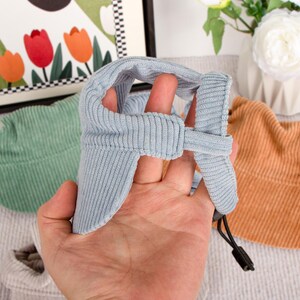 Pu&ograve; includere: Un cappello per animali domestici in velluto a coste azzurro con paraorecchie e sottogola regolabile. Altri cappelli in verde e arancione sono visibili sullo sfondo. I cappelli sono progettati per proteggere gli animali domestici dal sole.