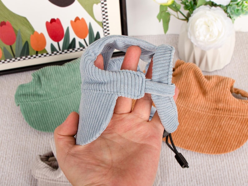 Pu&ograve; includere: Un cappello per animali domestici in velluto a coste azzurro con paraorecchie e sottogola regolabile. Altri cappelli in verde e arancione sono visibili sullo sfondo. I cappelli sono progettati per proteggere gli animali domestici dal sole.