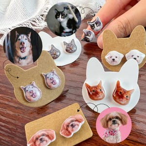 May include: A set of six pairs of custom pet portrait earrings. Each pair features a different pet, including a husky, a cat, a shih tzu, and a poodle. The earrings are displayed on brown and white cardboard cat-shaped earring display cards.