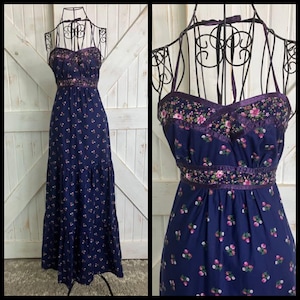 May include: A floor-length navy blue maxi dress featuring a small pink and white floral pattern, a sweetheart neckline, and thin halter-style straps. The bodice and waist are accented with a matching floral fabric band and dark purple ribbon trim.