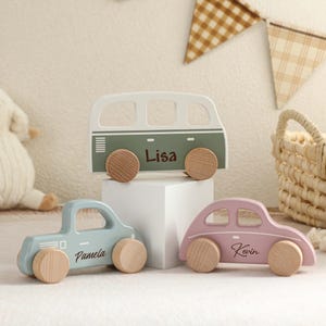 May include: Three wooden toy vehicles: a light blue pickup truck, a pink car, and a green and white camper van. Each has wooden wheels and a name printed on the side. The camper van says "Lisa". The pickup truck says "Pamela". The car says "Kevin".