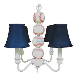 May include: A white baseball-themed chandelier with five black lampshades. The chandelier is decorated with four baseballs stacked on top of each other. The baseballs have red stitching and the words "Little League" are printed on one of the baseballs.