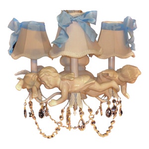 May include: A white chandelier with three light fixtures and blue ribbon shades. The chandelier is decorated with cherub figurines and crystal drops.